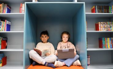 To elever sitter på biblioteket og leser, en i bok og en på læringsbrett.