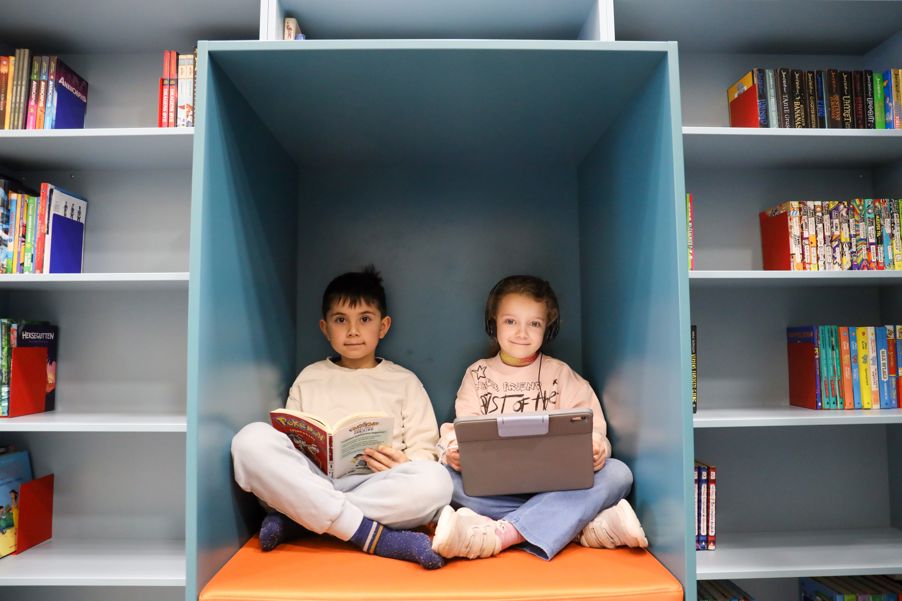 To elever sitter på biblioteket og leser, en i bok og en på læringsbrett.
