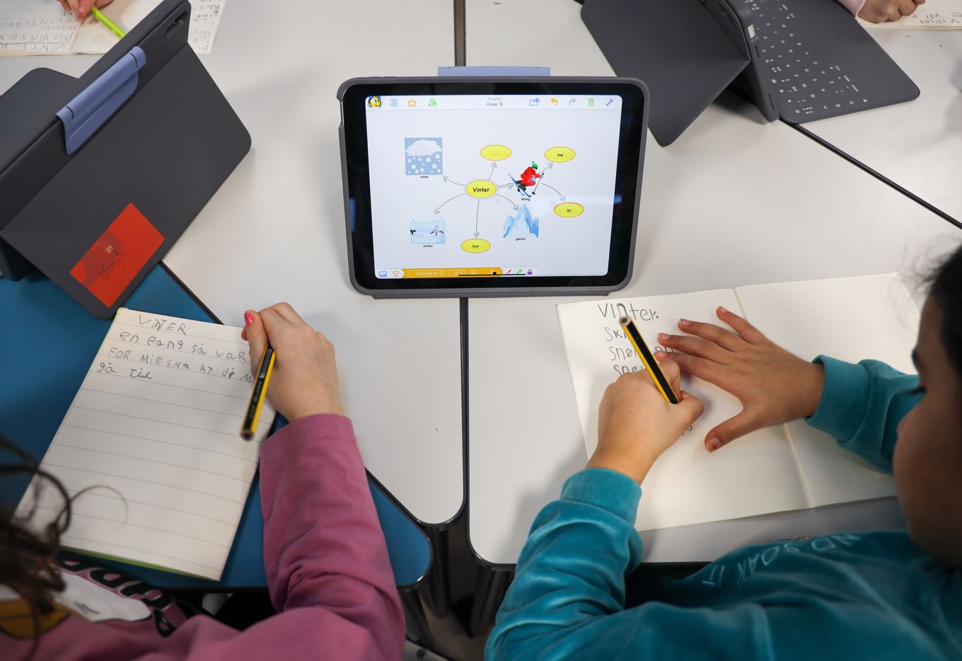 To elever som sitter ved et skrivebord og skriver i skriveboka med et digital tankekart foran seg. 