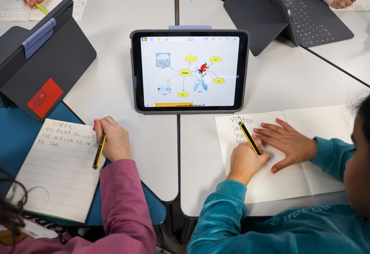 To elever som sitter ved et skrivebord og skriver i skriveboka med et digital tankekart foran seg. 